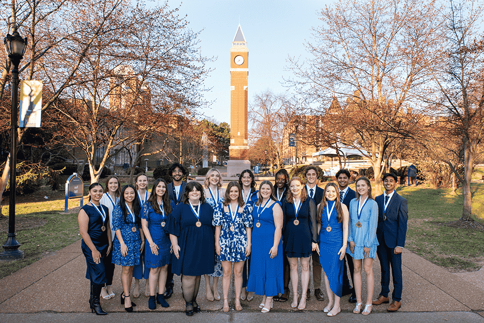 A group of 18 students wear medals around their neck with the clocktower over their heads in the background.